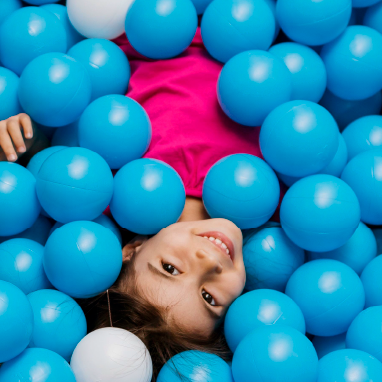 Miniatura de una niña mirando a cámara sonriendo tumbada sobre pelotas de juguete de color azul