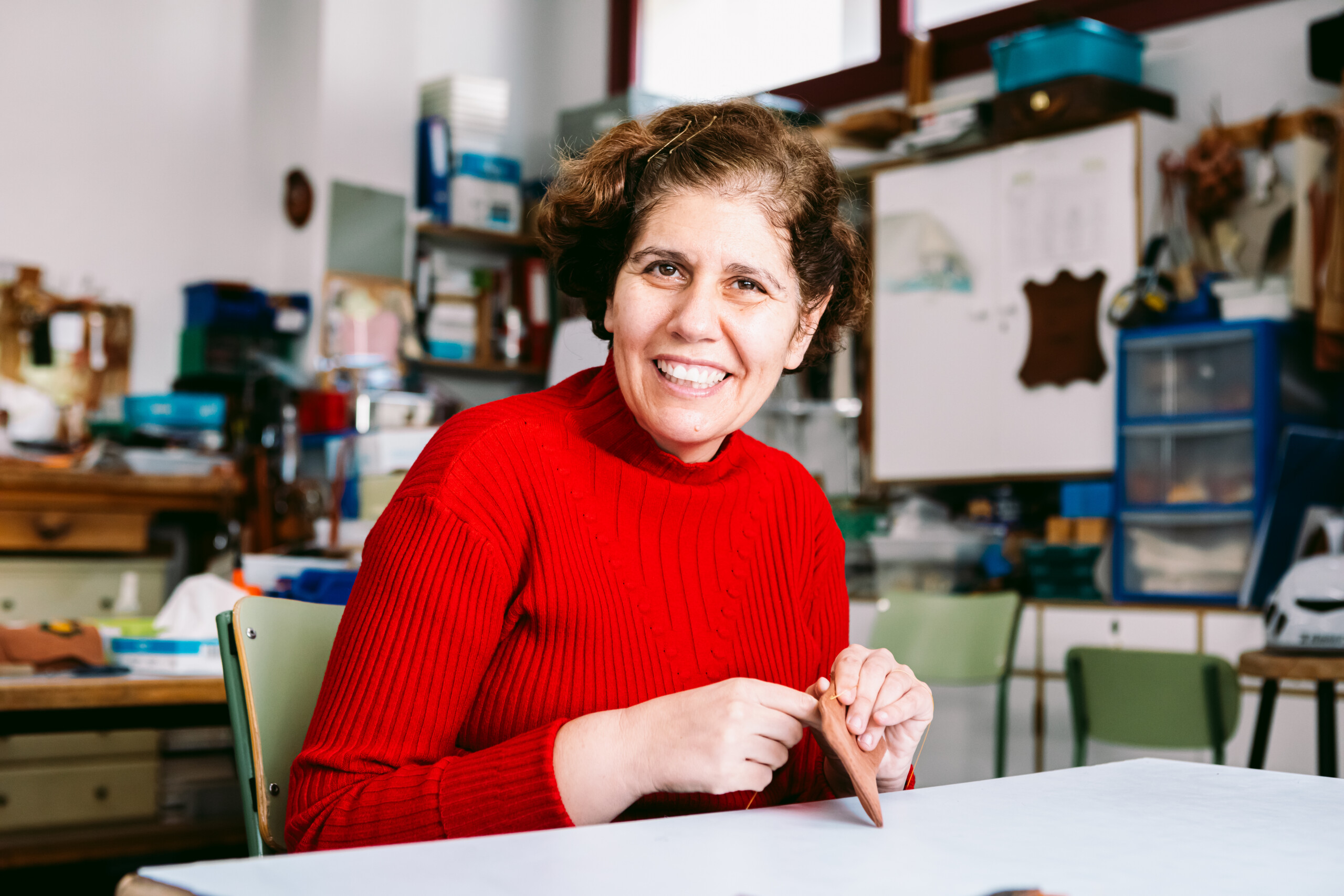 Mujer adulta trabajando sentada en un taller, cosiendo una pieza de cuero, y sonriendo a la cámara