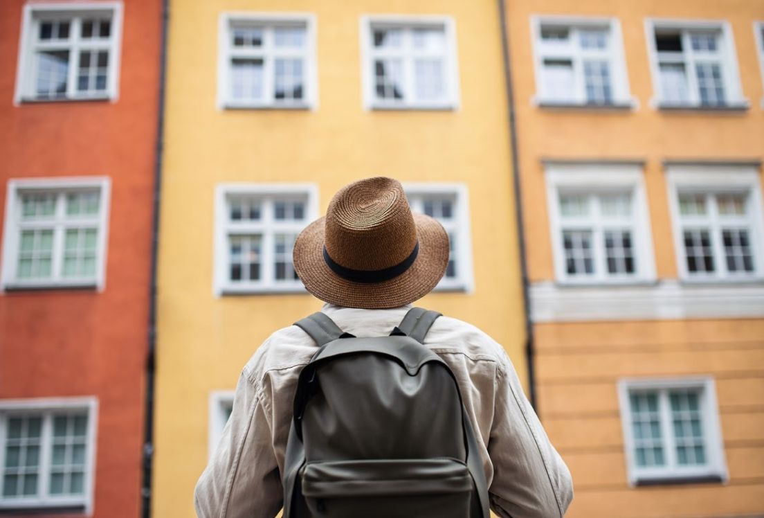 Fotografía en la que sale una persona de espalda con mochila y sombrero, observando una edificio.