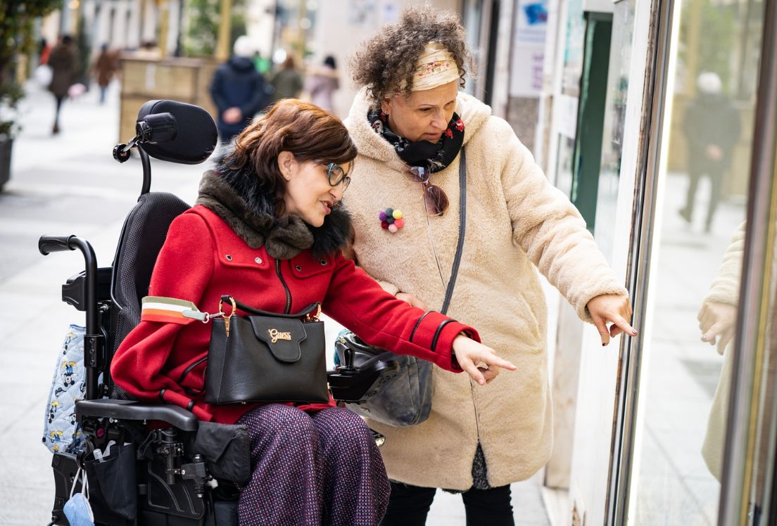 Una persona con discapacidad física observa y señala a un escaparate junto a otra mujer que está a su lado.