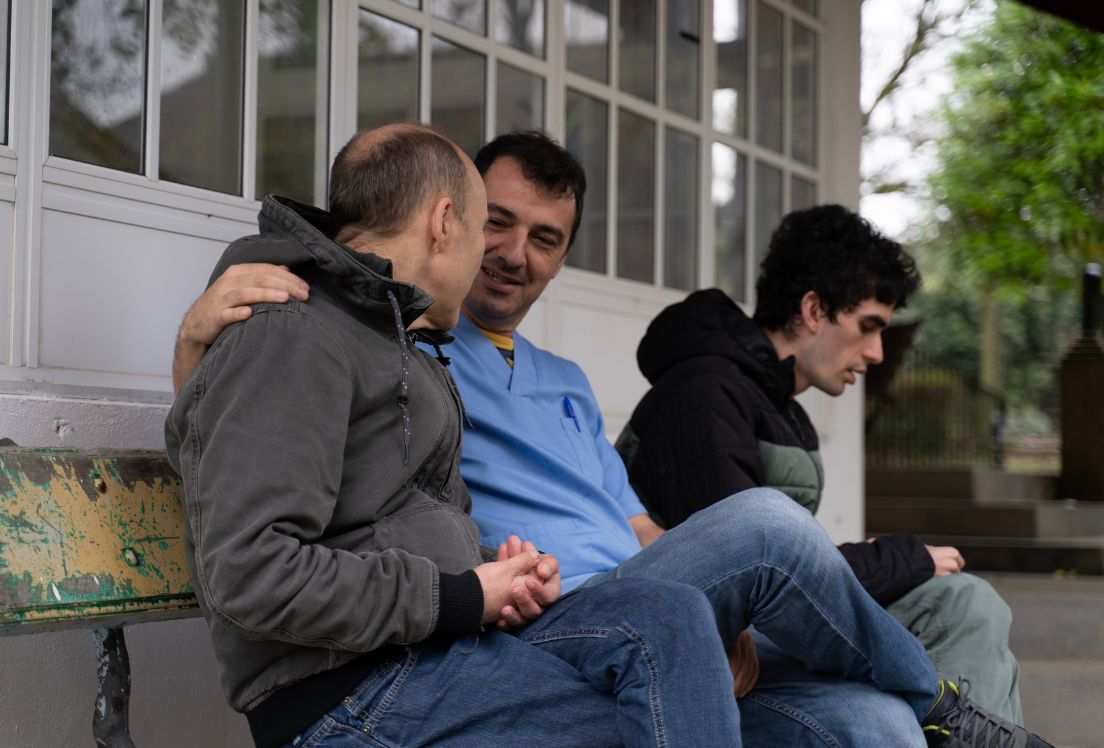 Fotografía de tres personas sentadas en un banco. el hombre que está en medio le pasa la mano por el hombro a una de ellos.