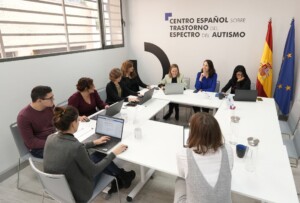 Fotografía de una reunión de trabajo: un grupo de personas mantienen una reunión de trabajo. en la pared del fondo el logo del Centro Español de Autismo. 