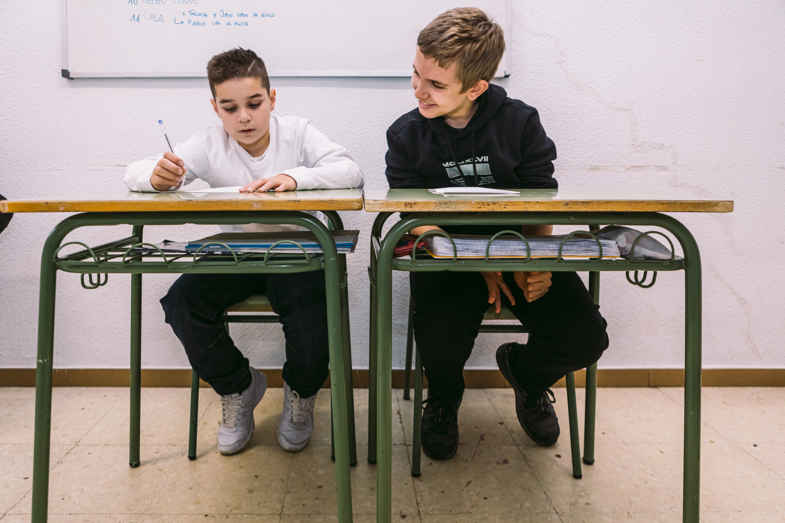 Dos niños sentados en unos pupitres contiguos con uno mirando lo que hace el otro