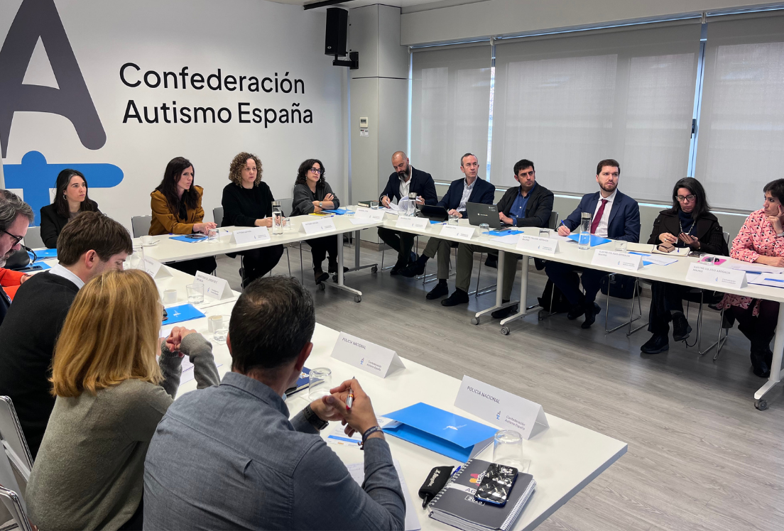 Un grupo de personas en un sala, sentados en una mesa dispuesta en forma de U con el logo de Autismo España al fondo.