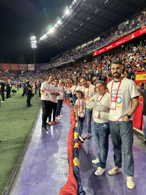 Un grupo de personas autistas, al pie de un campo de fútbol con camisetas blancas con un infinito multicolor