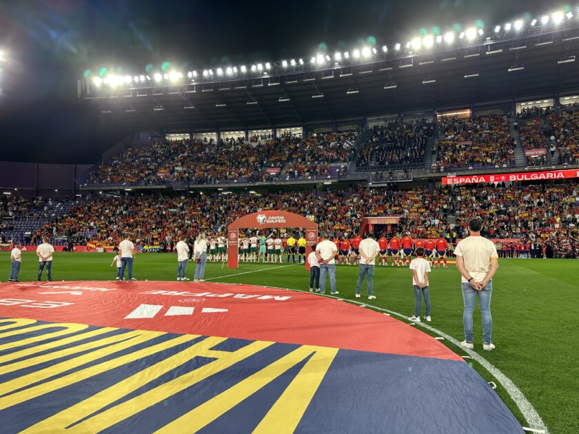 Vista de una lona posada en el césped de un campos de fútbol, con personas alrededor y el estadio de fondo.