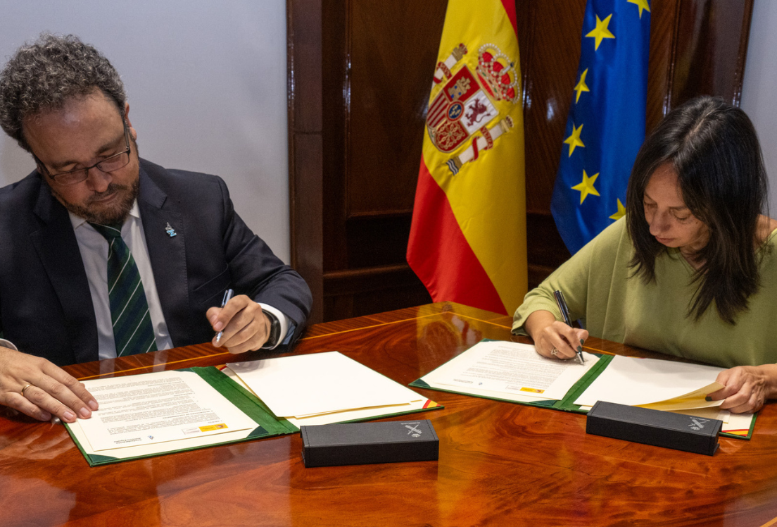 Un hombre y una mujer sentados a una mesa, firman un documento cada uno, con la bandera de España y la de la Unión Europea detrás