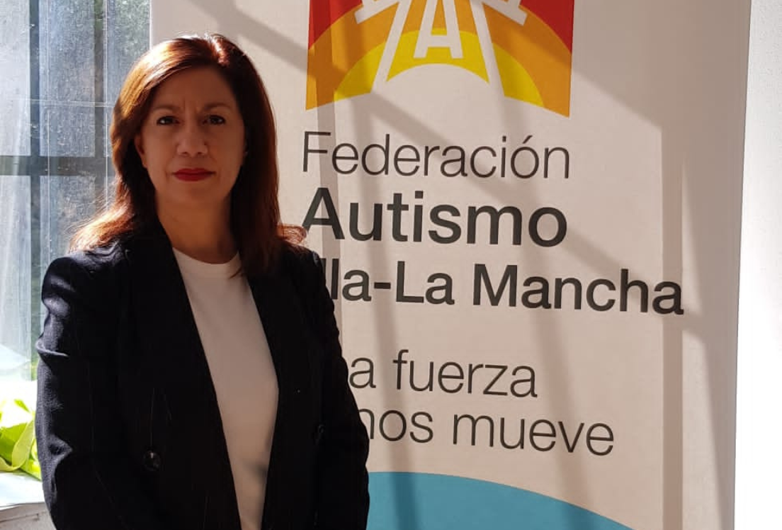 Medio plano de una mujer mirando a cámara. Detrás tiene un letrero de "federación Autismo Castilla-La Mancha"