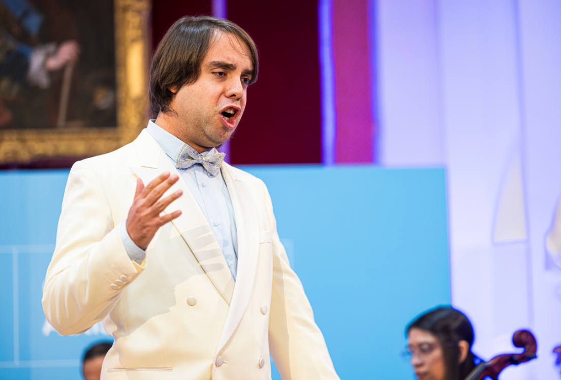 Medio plano de un joven que canta, vestido de blanco y con pajarita