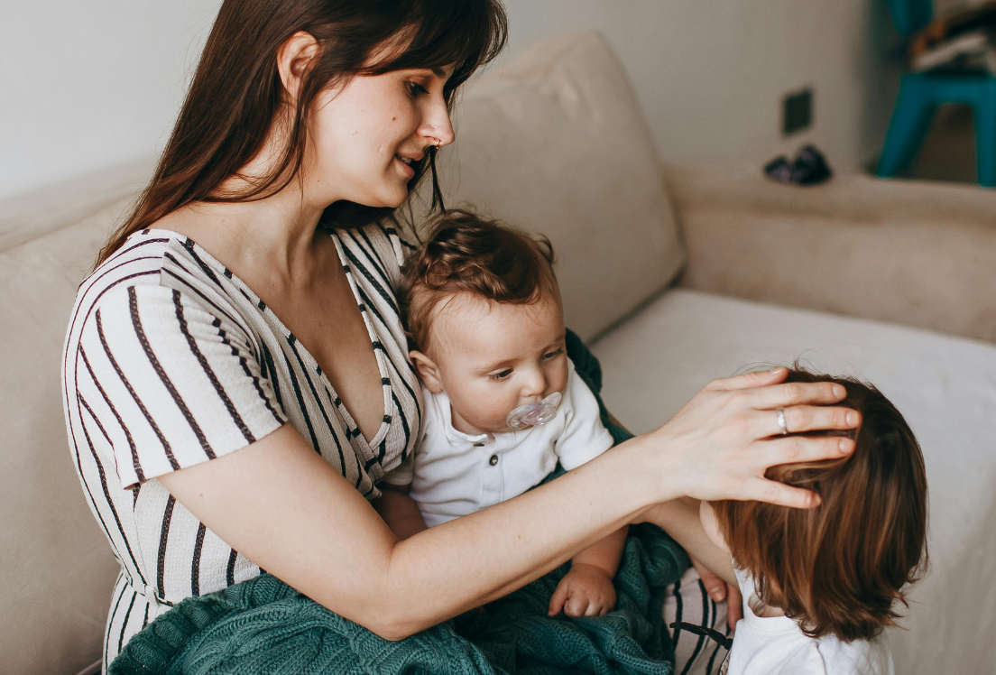 Mujer sentada con un bebé en brazos acaricia la cabeza de otro niño