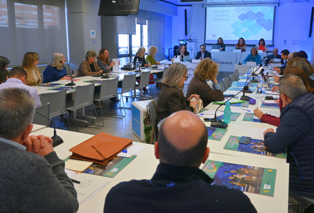 Un grupo de personas en una sala reunidas con una pantalla de fondo, en la asamblea general de Autismo España