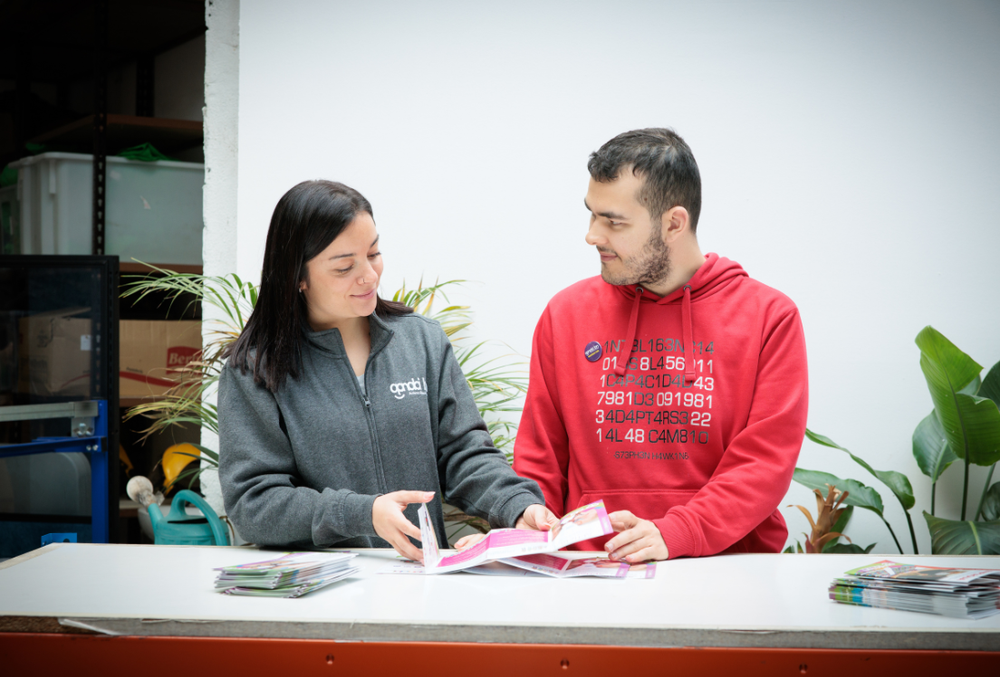 Un chico y una chica sonrien mientras doblan unos folletos de pie detrás de un mostrador