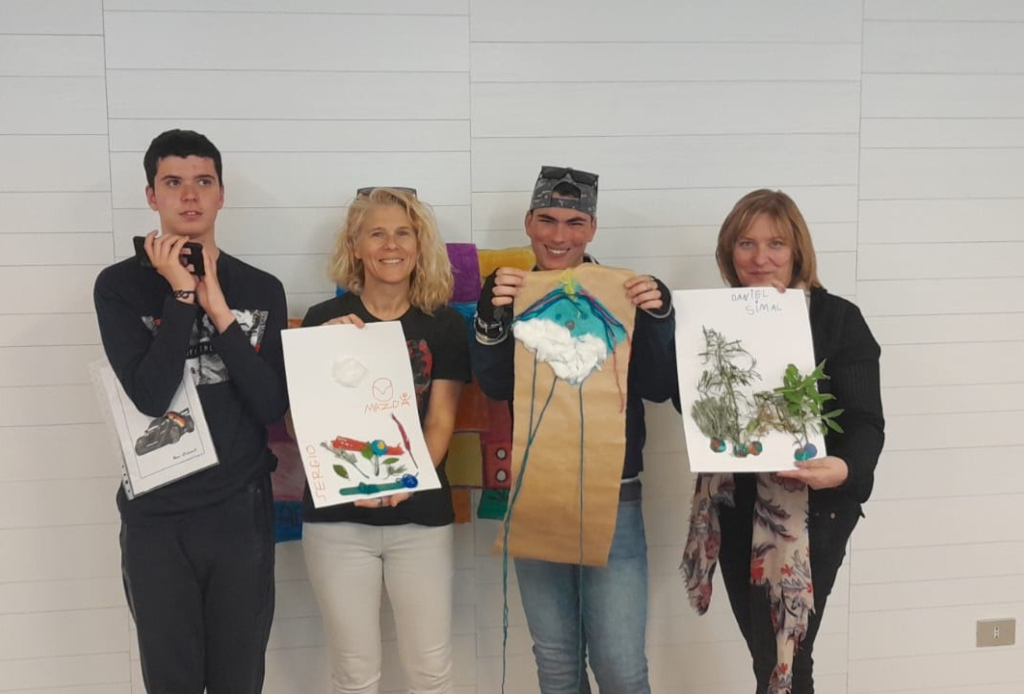 Cuatro personas de pie frente a una pared blanca sostienen trabajos artísticos relacionados con la naturaleza. De izquierda a derecha: un joven, una mujer, otro joven y una mujer